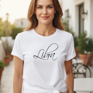 Könnte beinhalten: Weißes T-Shirt mit dem Wort "Libra" in einer stilvollen, kursiven Schrift. Das Shirt hat kurze Ärmel und eine lässige Passform. Das Design ist schlicht und elegant, ideal für alle, die sich für Astrologie und Tierkreiszeichen interessieren.