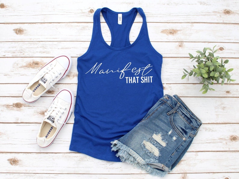 May include: A royal blue racerback tank top with the text "Manifest That Shit" in white script. The image also includes a pair of white canvas sneakers and denim shorts with distressed details.