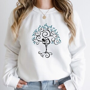 May include: White sweatshirt featuring a black tree design with a yoga pose figure incorporated into the trunk. The tree has teal leaves and swirling branches. The sweatshirt is a casual, comfortable style.