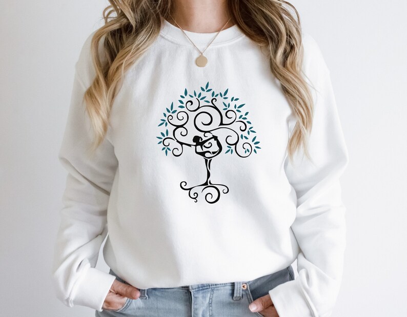 May include: White sweatshirt featuring a black tree design with a yoga pose figure incorporated into the trunk. The tree has teal leaves and swirling branches. The sweatshirt is a casual, comfortable style.