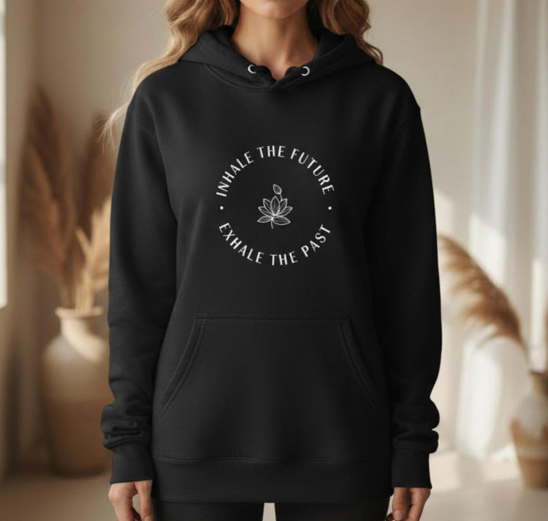 May include: Black hoodie with a front pocket and drawstring hood. The hoodie features a white circular graphic with the text "INHALE THE FUTURE, EXHALE THE PAST" and a lotus flower illustration. The hoodie is made of a soft material.