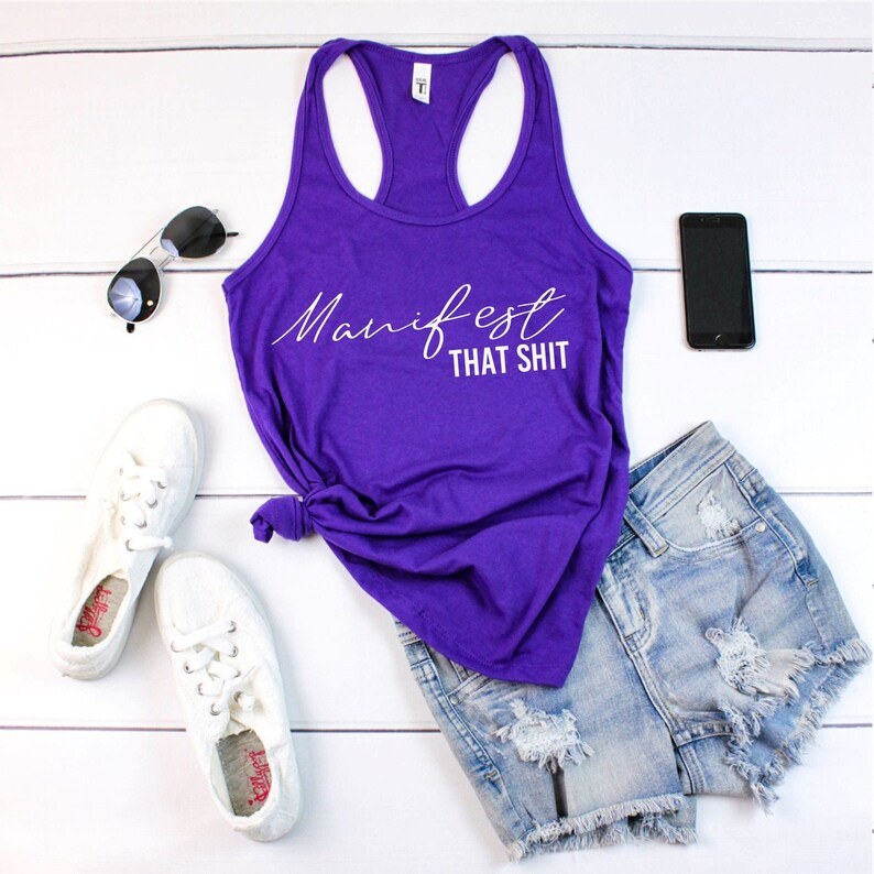 May include: A purple tank top with the text "Manifest That Shit" in white script is paired with denim shorts, white sneakers, sunglasses, and a smartphone. The casual outfit is arranged on a white wooden surface.