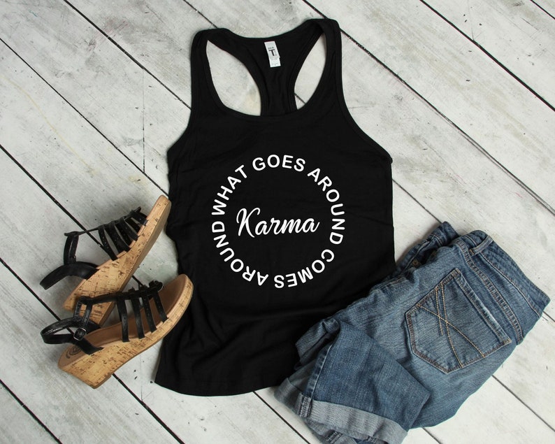 May include: Black racerback tank top with the text "Karma" in a white circular design. The tank top is paired with denim shorts and black sandals with cork soles. The items are arranged on a light wood-grain surface.