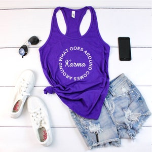 May include: A purple tank top with the text "Karma, what goes around comes around" in a circular design. The image also includes denim shorts, white sneakers, sunglasses, and a black smartphone, all arranged on a white wooden surface.