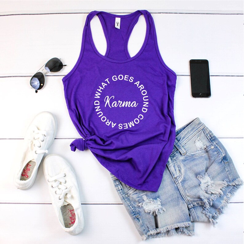 May include: A purple tank top with the text "Karma, what goes around comes around" in a circular design. The image also includes denim shorts, white sneakers, sunglasses, and a black smartphone, all arranged on a white wooden surface.