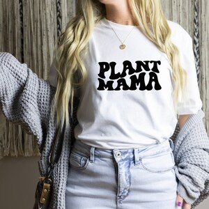 K&ouml;nnte beinhalten: Wei&szlig;es T-Shirt mit dem Text "PLANT MAMA" in schwarzer, retroartiger Schrift. Das Shirt wird mit hellblauen Jeans und einer grauen Strickjacke kombiniert. Das Gesamtbild deutet auf ein l&auml;ssiges Thema f&uuml;r Pflanzenliebhaber hin.
