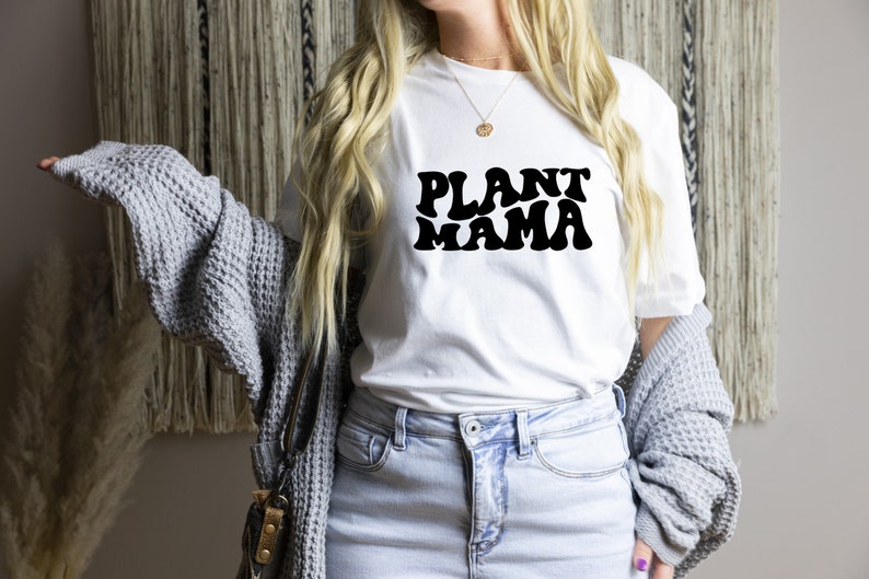 K&ouml;nnte beinhalten: Wei&szlig;es T-Shirt mit dem Text "PLANT MAMA" in schwarzer, retroartiger Schrift. Das Shirt wird mit hellblauen Jeans und einer grauen Strickjacke kombiniert. Das Gesamtbild deutet auf ein l&auml;ssiges Thema f&uuml;r Pflanzenliebhaber hin.