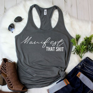 May include: A dark gray racerback tank top with the words "Manifest THAT SHIT" in white script. The tank top is styled with sunglasses, brown ankle boots, and blue jeans.