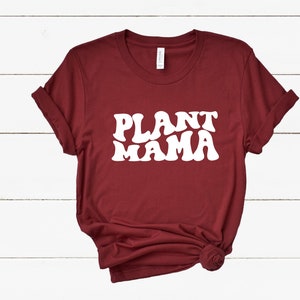 K&ouml;nnte beinhalten: Ein weinrotes T-Shirt mit dem Schriftzug "PLANT MAMA" in gro&szlig;en, wei&szlig;en Buchstaben im Retro-Stil. Die &Auml;rmel sind hochgekrempelt und der Saum ist geknotet. Das T-Shirt ist aus einem weichen Material.