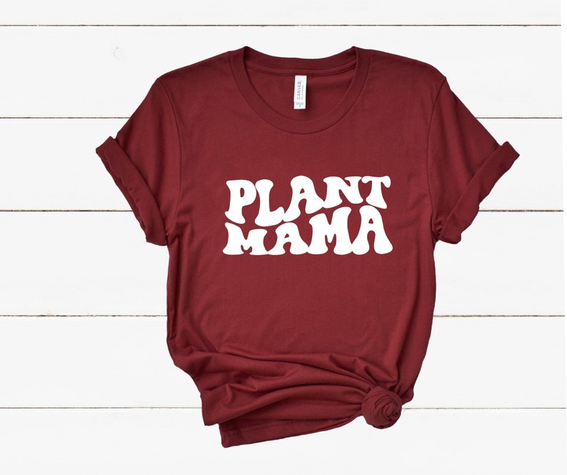 K&ouml;nnte beinhalten: Ein weinrotes T-Shirt mit dem Schriftzug "PLANT MAMA" in gro&szlig;en, wei&szlig;en Buchstaben im Retro-Stil. Die &Auml;rmel sind hochgekrempelt und der Saum ist geknotet. Das T-Shirt ist aus einem weichen Material.