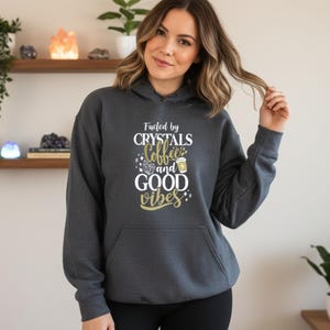 May include: Dark gray hoodie with a front pocket and a drawstring hood. The hoodie features the text "Fueled by Crystals, Coffee, and Good Vibes" in white and gold lettering. The design includes a coffee cup and crystal illustrations.