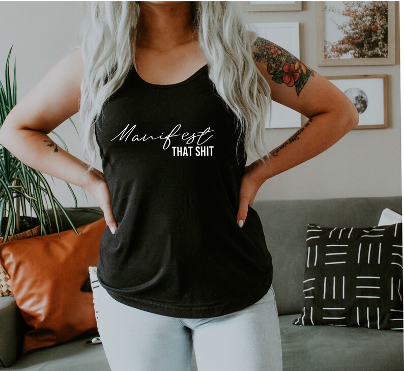 May include: Black tank top with the white text "Manifest That Shit". The top is a sleeveless style, made of a soft material. The text is in a cursive font. The tank top is worn with light blue jeans.