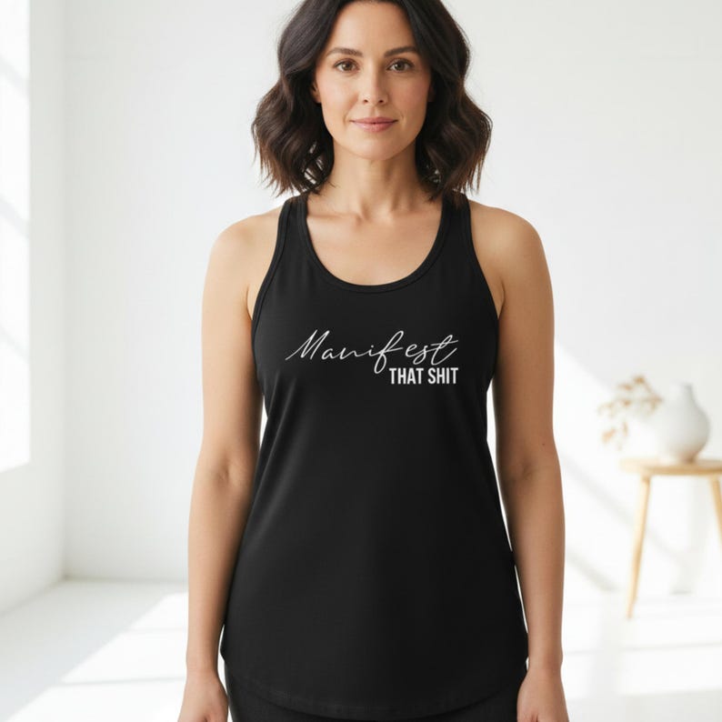 May include: Black tank top with the white text "Manifest That Shit" in a cursive font. The tank top has a racerback design and a scoop neckline. The image shows a woman wearing the tank top.