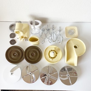 May include: A set of kitchen appliance attachments, including a set of beaters, a set of blades, a set of grater attachments, and a set of mixing bowls. The attachments are all in white and beige.