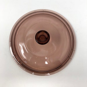 May include: A round, transparent, light purple glass lid with a central knob. The lid features a series of concentric ridges radiating from the center, creating a textured pattern. The knob is a darker shade of purple.