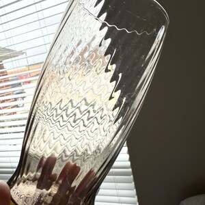 May include: A tall, clear glass tumbler featuring a distinct rippled, wavy texture throughout its surface. The glass has a slightly flared rim and a tapered base, with a smooth, transparent finish that catches light to highlight the undulating pattern.