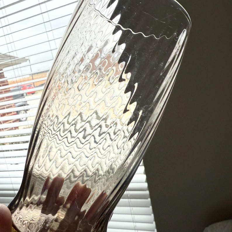 May include: A tall, clear glass tumbler featuring a distinct rippled, wavy texture throughout its surface. The glass has a slightly flared rim and a tapered base, with a smooth, transparent finish that catches light to highlight the undulating pattern.