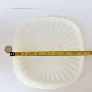 May include: A square, off-white plastic lid with a sunburst pattern of raised ridges radiating from the center. A yellow Stanley measuring tape is placed across the lid, showing a width of approximately 8.5 inches.