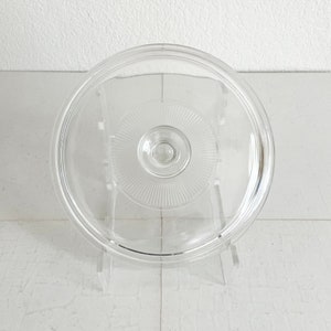 May include: A clear glass lid with a circular design. The lid features a central knob and a pattern of radiating lines. The lid is displayed on a clear stand against a white background, showcasing its transparency and design.