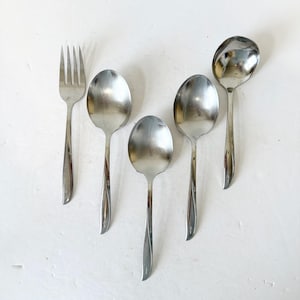 Puede incluir: Un conjunto de cubiertos antiguos, que incluye cuatro cucharas y un tenedor, dispuestos sobre una superficie blanca. Las cucharas tienen cuencos ovalados y mangos delgados. El tenedor tiene cuatro púas. La cubertería es de metal brillante color plata.