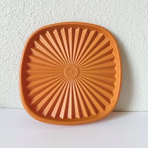 May include: A square, orange plastic plate with rounded corners, featuring a sunburst pattern of raised, radiating ridges that extend from a central circular hub. The design creates a symmetrical, pleated effect across the entire surface.