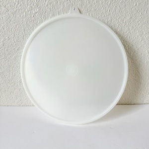 Puede incluir: Una tapa de plástico blanca y redonda con un pequeño asa en la parte superior. La tapa es ligeramente translúcida y tiene una superficie lisa. La tapa está diseñada para encajar en un recipiente. Es un artículo de almacenamiento de cocina.