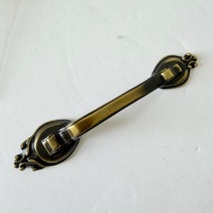 Könnte beinhalten: Ein Möbelgriff im antiken Stil mit dunkelbronzenem Finish und goldenen Akzenten. Der Griff hat dekorative ovale Endplatten und einen geraden Stab. Hardware für Möbelrestaurierung oder Neubauten.