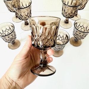 May include: A collection of vintage brown-tinted glass goblets. Each goblet features a decorative diamond pattern on the bowl and a stemmed base. The glasses are arranged on a white surface, with one held in a hand.