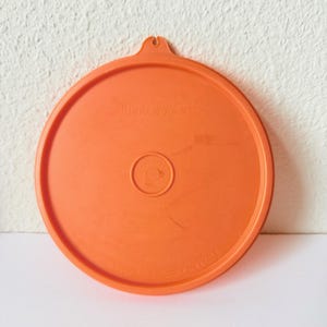 May include: A round, orange plastic Tupperware lid featuring a small tab at the top for easy removal. The center has a circular indentation, and the surface is embossed with the brand name Tupperware and the product code 227-48.