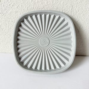 May include: A light grey, square Tupperware lid with a sunburst design. The lid has rounded corners and a circular indentation in the centre. The design features radiating lines from the centre to the edge.
