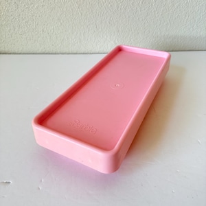 May include: A rectangular, light pink Barbie-branded tray. The tray has a raised edge and a slightly recessed interior. The Barbie logo is embossed on the lower left corner. The tray is made of plastic and is likely a vintage toy accessory.