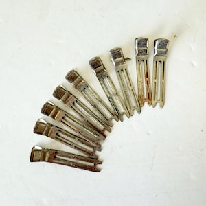 May include: A collection of vintage metal hair clips arranged in a semi-circle. The clips are silver-toned with a rectangular top and a slotted design. Some clips show signs of age and wear, with a slightly tarnished appearance.