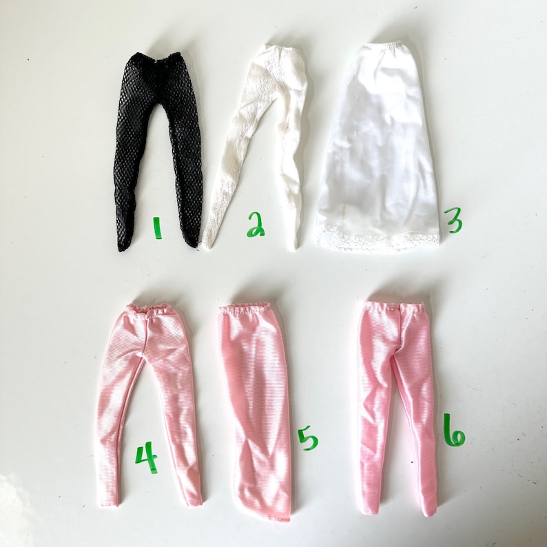 Op de afbeelding: Zes verschillende kledingstukken voor poppen, waaronder zwarte kousen met visnet, witte kousen met kant, een witte slip, een roze satijnen broek en twee paar roze satijnen broeken.