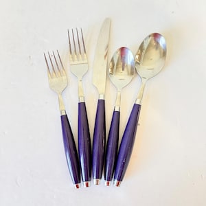 May include: A set of five stainless steel utensils: two forks, a knife, and two spoons. Each piece has a dark purple handle with a silver band and a red tip. The utensils are arranged on a white surface.