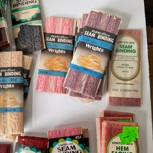 May include: Assortment of vintage seam binding and hem facing rolls in various colors, including pink, mauve, and beige. The packaging features the brands J. & P. Coats and Wrights, with text indicating nylon material and measurements in yards.