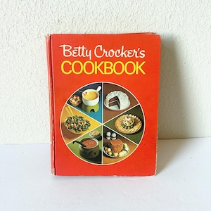 Könnte beinhalten: Ein altes Betty Crocker's COOKBOOK mit einem leuchtend orangefarbenen Einband. Die Vorderseite zeigt eine kreisförmige Grafik, die in sechs Abschnitte unterteilt ist, von denen jeder ein anderes Gericht zeigt, darunter Kuchen, Fondue, Salat und Suppe.