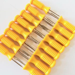 May include: A set of twelve corn cob holders with bright yellow plastic handles featuring a ribbed, cylindrical grip design and two sharp, parallel silver-coloured metal prongs extending from each handle for securing food items during consumption.