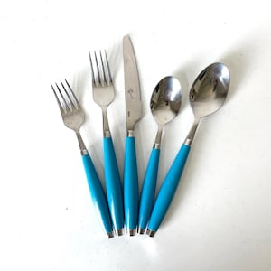 May include: A set of five pieces of silverware: two forks, a knife, and two spoons. The handles are a bright turquoise blue, and the metal is silver. The knife has a small floral design on the blade. The silverware is arranged on a white surface.