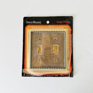 Puede incluir: Una placa de pared vintage de Deco Room para dos interruptores de luz. La placa tiene un patrón de vetas de madera y un borde decorativo de color plateado. El embalaje incluye el texto "Wall Plate" y "American Tack & Hardware Co., Inc., Monsey, N.Y. 10952".