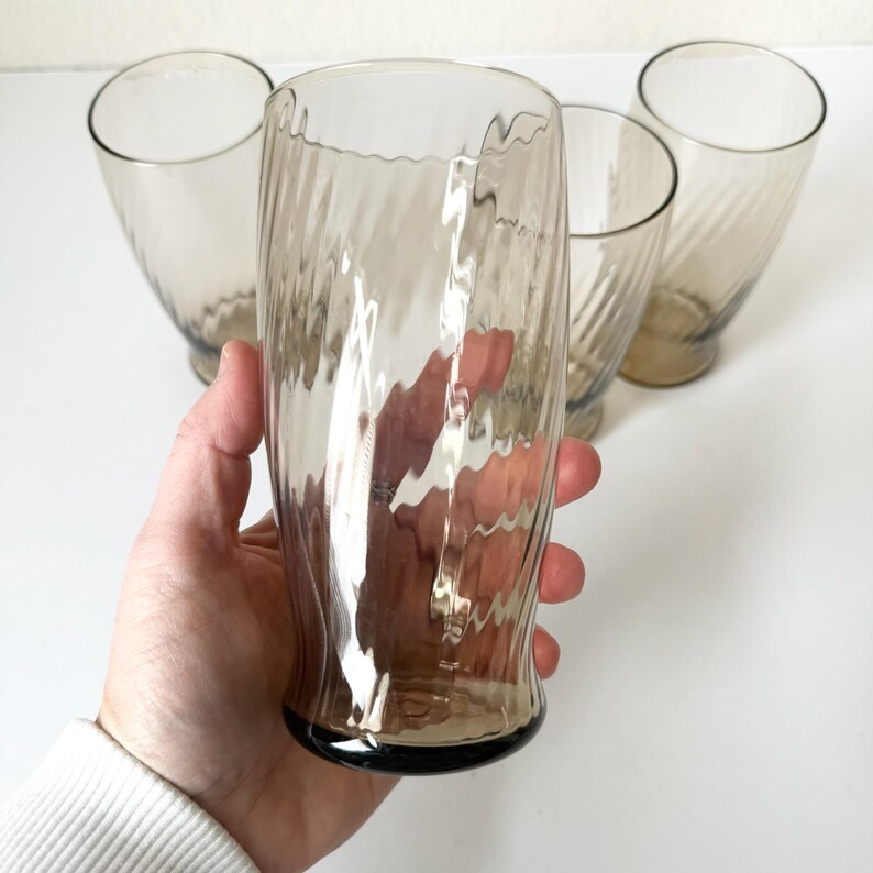 May include: A set of four tall drinking glasses made of translucent smoke-colored glass. Each glass features a distinct vertical ribbed or fluted texture along the exterior surface, creating a wavy, light-reflecting pattern throughout the body.