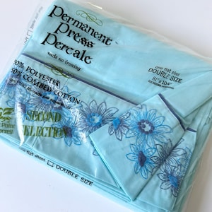 以下が含まれることがあります： プラスチック包装に入った水色のフラットシーツ。Permanent Press Percale、ポリエステル50%、コーマ綿50%、ダブルサイズ205.7cm x 264.2cmと記載。縁には青と濃い青の刺繍の花柄が施されている。