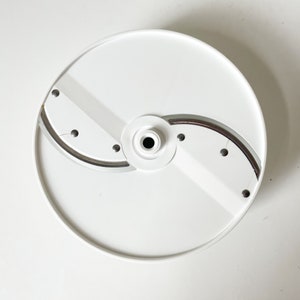 May include: White plastic food processor blade with three silver blades and a central hub. The blade is designed for slicing and shredding.