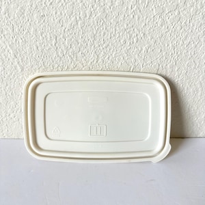 May include: A rectangular, off-white plastic food storage container lid with rounded corners. The lid features a raised rim, a central recessed panel with embossed text, a recycling symbol, and a small icon depicting two vertical lines.
