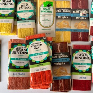 May include: A collection of vintage seam binding and hem facing rolls in various colors. The packages display text such as "Stretch Lace Seam Binding" and "J. & P. Coats." Colors include yellow, peach, red, and orange. Each roll is labeled with the yardage.