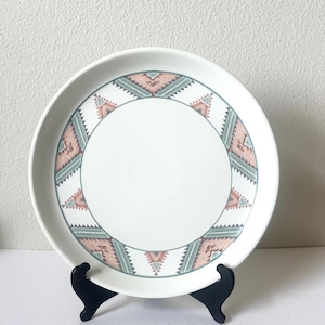 May include: A round white ceramic plate featuring a decorative border with a repeating geometric pattern in muted shades of dusty pink, sage green, and dark gray. The center of the plate is plain white, and the rim is smooth and circular.