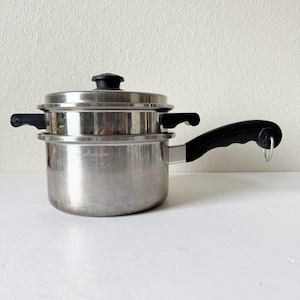 May include: Stainless steel double boiler with a lid and black handles. The pot has a long handle on the side and two smaller handles on the sides of the upper pot. The lid has a black knob.