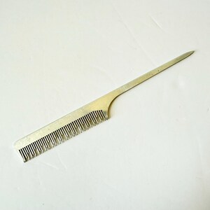 May include: A silver-colored metal comb with a long, pointed handle. The comb has fine teeth along one edge and is designed for hair styling. The comb is made of a shiny, metallic material.