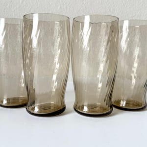 May include: A set of four tall drinking glasses made of translucent smoke-colored glass. Each glass features a distinct vertical ribbed or fluted texture that creates a swirling pattern along the cylindrical body, with a smooth, clear rim.