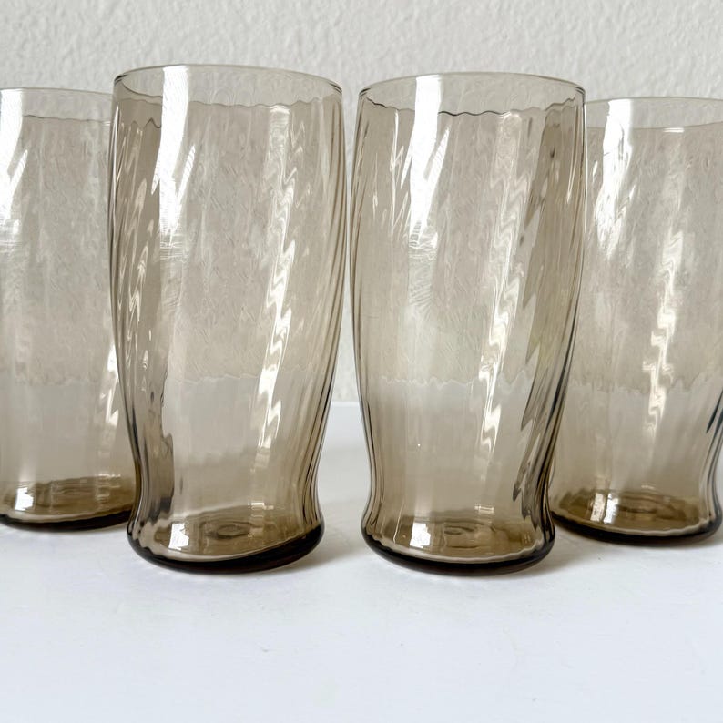 May include: A set of four tall drinking glasses made of translucent smoke-colored glass. Each glass features a distinct vertical ribbed or fluted texture that creates a swirling pattern along the cylindrical body, with a smooth, clear rim.