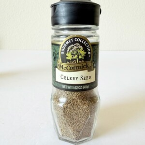 May include: A clear glass jar of McCormick Celery Seed with a black plastic lid. The label features the words "Gourmet Collection" and "Celery Seed". The net weight is 1.62 oz (45g). The jar is filled with brown celery seeds.